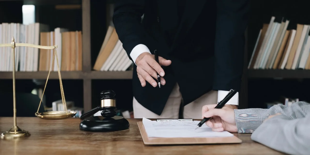 Advogado e cliente assinam um contrato em uma mesa de escritório, com a balança da justiça e um martelo de juiz ao lado