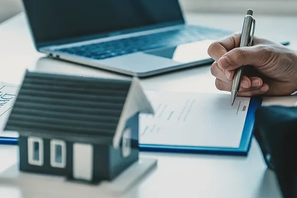 Pessoa assina um contrato de compra e venda ao lado de uma miniatura de casa e de um notebook