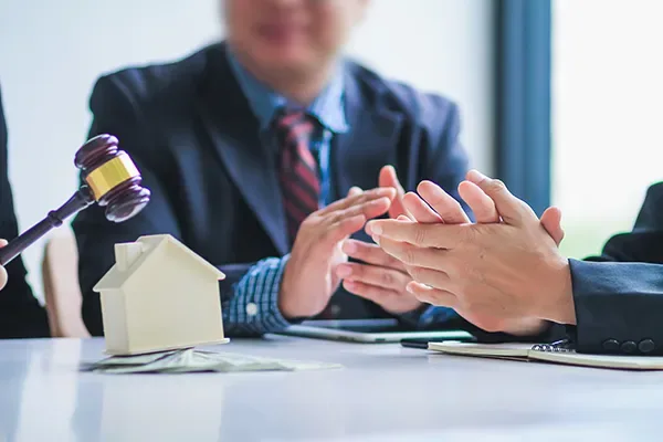 Martelo de leilão e miniatura de casa sobre a mesa enquanto duas pessoas fecham negócio com um aperto de mãos